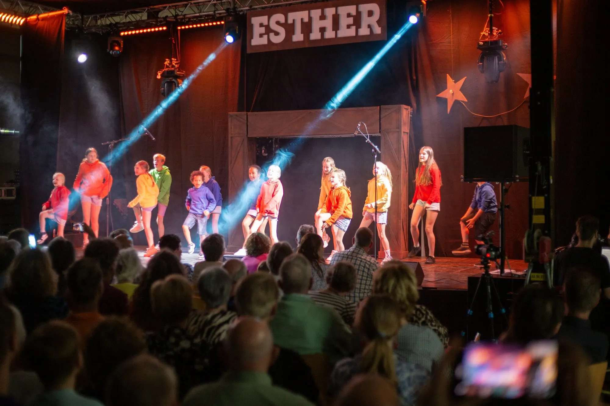 Kinderen in kleurrijke kostuums op het podium van Esther, met lichtbundels door de rook en een volle zaal