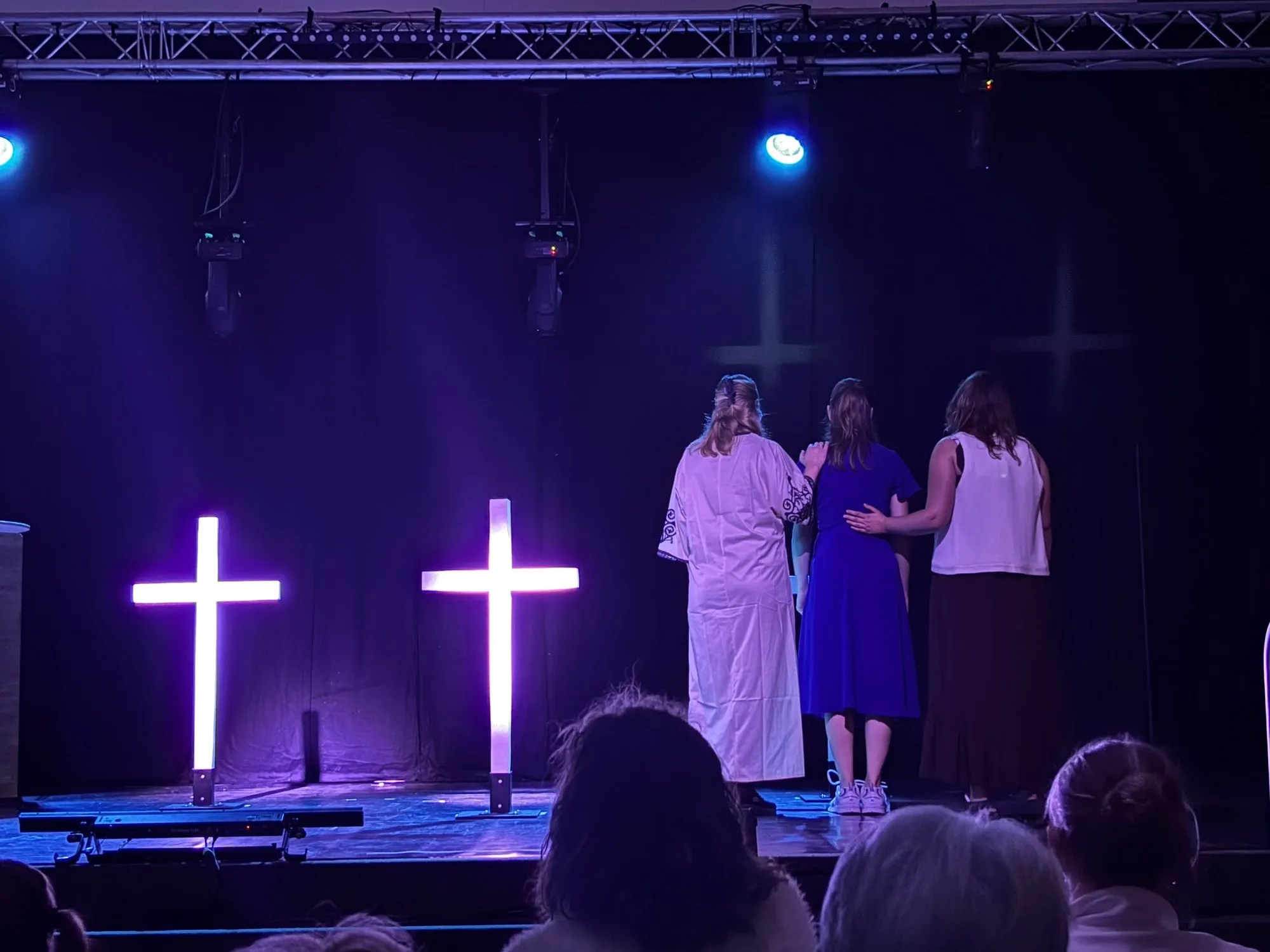 Twee verlichte kruizen in paars licht, met spelers in lange gewaden op het podium van Ruth